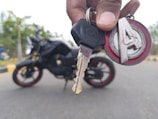 A close-up of a hand holding a motorcycle key with a branded keychain. In the background, there is a motorcycle with red and black detailing, situated on a road with blurred trees in the distance.