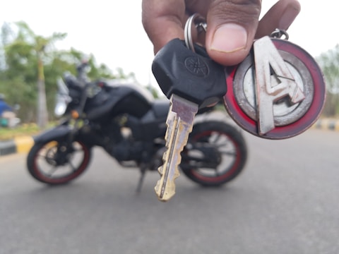 A close-up of a hand holding a motorcycle key with a branded keychain. In the background, there is a motorcycle with red and black detailing, situated on a road with blurred trees in the distance.