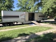 A large moving truck is parked on a residential street with its back doors open and a ramp leading down to the sidewalk. Boxes and furniture can be seen inside the truck, and a person is partially visible inside, seemingly organizing or unloading items. The surrounding area has lush green trees and grass, suggesting it’s a well-kept neighborhood.