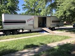 A large moving truck is parked on a residential street with its back doors open and a ramp leading down to the sidewalk. Boxes and furniture can be seen inside the truck, and a person is partially visible inside, seemingly organizing or unloading items. The surrounding area has lush green trees and grass, suggesting it’s a well-kept neighborhood.