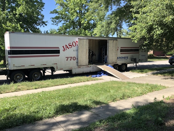 A large moving truck is parked on a residential street with its back doors open and a ramp leading down to the sidewalk. Boxes and furniture can be seen inside the truck, and a person is partially visible inside, seemingly organizing or unloading items. The surrounding area has lush green trees and grass, suggesting it’s a well-kept neighborhood.