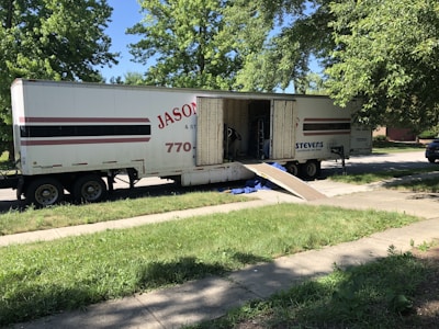 A large moving truck is parked on a residential street with its back doors open and a ramp leading down to the sidewalk. Boxes and furniture can be seen inside the truck, and a person is partially visible inside, seemingly organizing or unloading items. The surrounding area has lush green trees and grass, suggesting it’s a well-kept neighborhood.