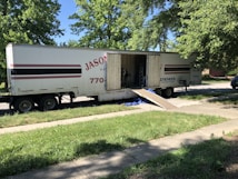 A large moving truck is parked on a residential street with its back doors open and a ramp leading down to the sidewalk. Boxes and furniture can be seen inside the truck, and a person is partially visible inside, seemingly organizing or unloading items. The surrounding area has lush green trees and grass, suggesting it&rsquo;s a well-kept neighborhood.