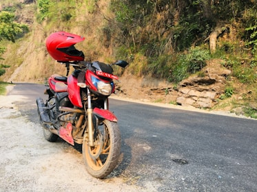 A red motorcycle is parked on the side of a paved road with a red helmet placed on its seat. The road stretches alongside a hillside covered with grass and shrubs. There are patches of sunlight filtering through the trees, creating a calm and serene atmosphere.