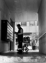 A person stands in a corridor, facing a wall of mailboxes, holding some paper or mail items. Behind them is a cart filled with packages. The area is partially covered, with recessed lighting on the ceiling, and the bright outside area is visible beyond the person.