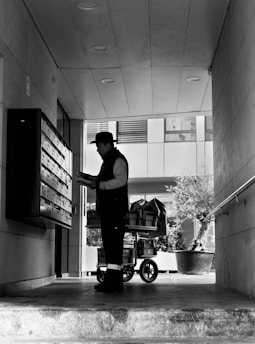 A person stands in a corridor, facing a wall of mailboxes, holding some paper or mail items. Behind them is a cart filled with packages. The area is partially covered, with recessed lighting on the ceiling, and the bright outside area is visible beyond the person.