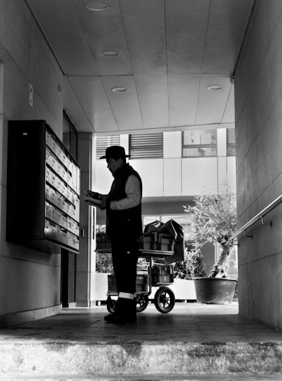 A person stands in a corridor, facing a wall of mailboxes, holding some paper or mail items. Behind them is a cart filled with packages. The area is partially covered, with recessed lighting on the ceiling, and the bright outside area is visible beyond the person.
