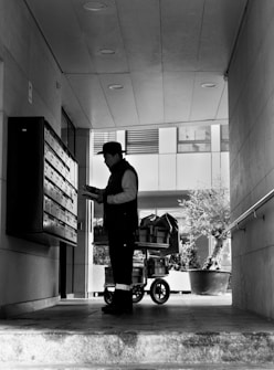 A person stands in a corridor, facing a wall of mailboxes, holding some paper or mail items. Behind them is a cart filled with packages. The area is partially covered, with recessed lighting on the ceiling, and the bright outside area is visible beyond the person.
