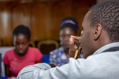 A group of individuals is gathered in what appears to be a social or educational setting. In the foreground, a man holding a pencil is visible, while in the background, two women are seated, with one wearing a red shirt. The background is blurred, emphasizing the person in the foreground.