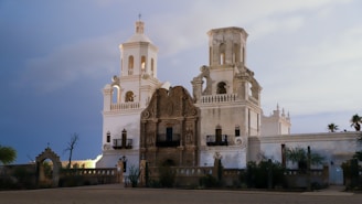 A historic mission building featuring two bell towers with intricate architectural details. The facade combines white and earthy tones, with a decorative central panel between the towers. The surrounding landscape includes desert vegetation and a decorative fence, set against a sky transitioning between day and night.