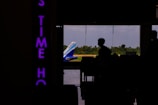 A silhouette of a person is visible inside an airport terminal, with the tail of an IndiGo airplane seen through large glass windows. Beyond the plane, there is a view of green trees and a blue sky. On the left, there is a vertically oriented neon sign with partially visible purple letters.
