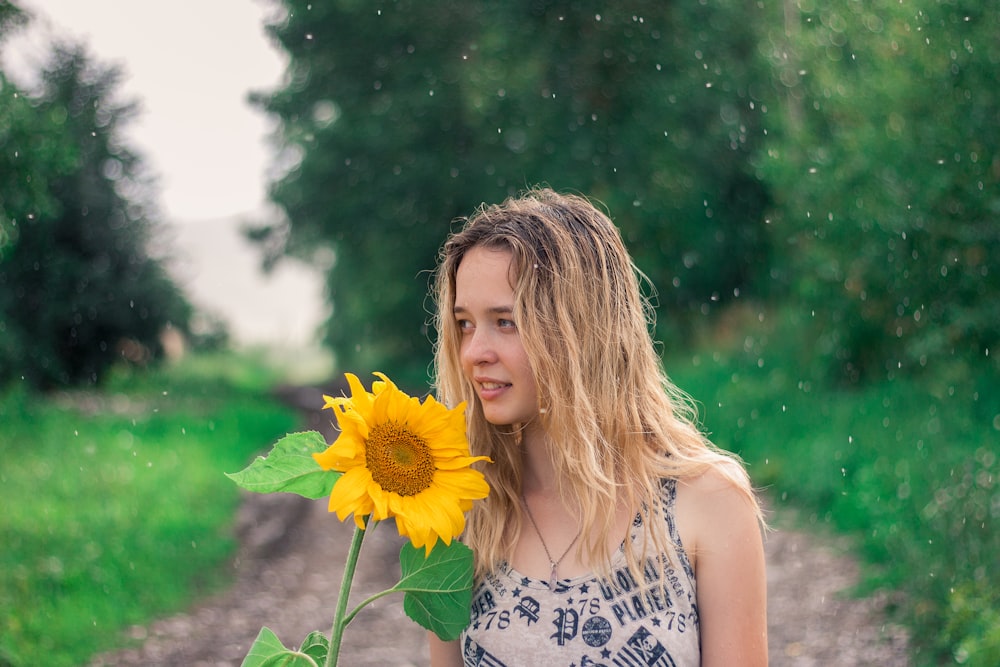 selective focus photography of woman standing near outdoor while holding sunflower during daytime