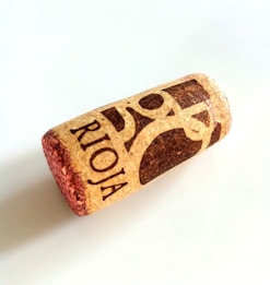 A wine cork with the word 'RIOJA' printed on it in bold letters along with a partial graphic design in dark ink. The cork has a natural appearance with a textured surface and is lying on a flat, light-colored background.