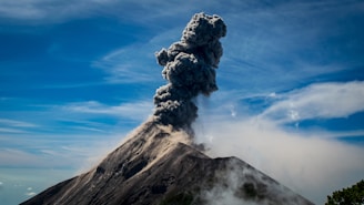 A volcano erupts, emitting a large cloud of dark ash and smoke into the sky. The cone-shaped mountain is surrounded by smoke as the eruption continues. The sky is bright blue with some wispy clouds scattered across it.