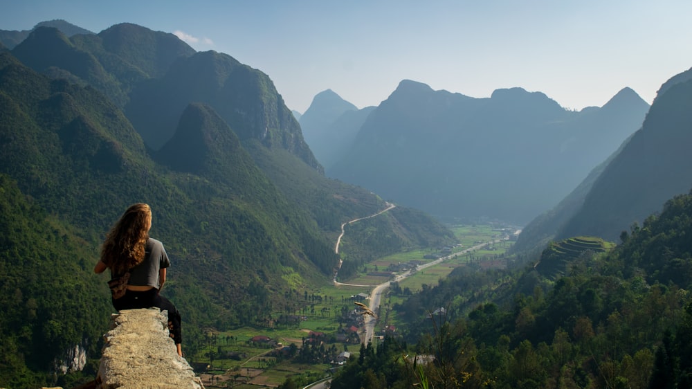 Photographie de femme assise sur une formation rocheuse faisant face à une chaîne de montagnes pendant la journée