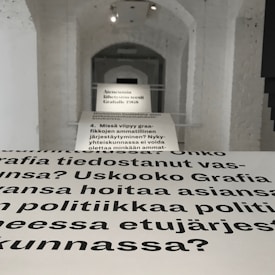 Text is displayed on a series of white panels arranged in a corridor with an arched ceiling. The text is in a bold, black font and appears to discuss professional organization and social responsibility, based on Finnish language text. The background consists of white brick walls and a white ceiling illuminated by recessed lighting.