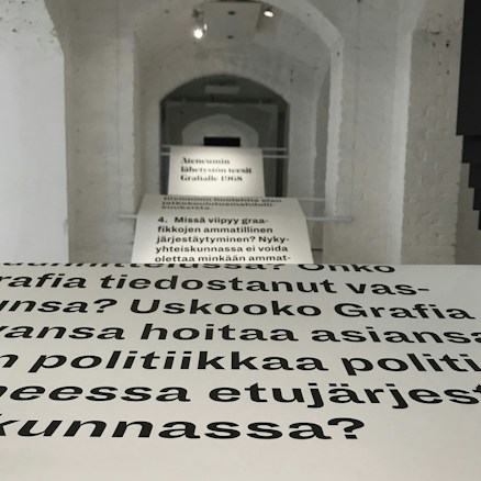 Text is displayed on a series of white panels arranged in a corridor with an arched ceiling. The text is in a bold, black font and appears to discuss professional organization and social responsibility, based on Finnish language text. The background consists of white brick walls and a white ceiling illuminated by recessed lighting.