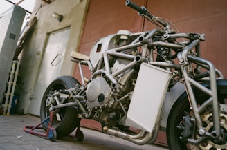 A stripped-down motorcycle without its bodywork, prominently displaying its complex metal frame and engine. The bike is placed on a stand in a garage or workshop setting, characterized by industrial elements like a concrete floor and metal door.