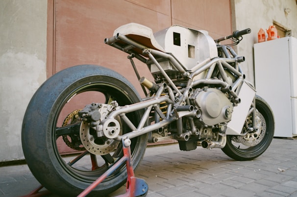 A custom-built motorcycle frame with an exposed skeletal structure is parked indoors. The metal framework showcases intricate welds and an unfinished appearance, highlighting the mechanical components like the engine and suspension. The rear tire is large and slick, indicating it may be designed for racing or high performance.