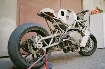 A custom-built motorcycle frame with an exposed skeletal structure is parked indoors. The metal framework showcases intricate welds and an unfinished appearance, highlighting the mechanical components like the engine and suspension. The rear tire is large and slick, indicating it may be designed for racing or high performance.