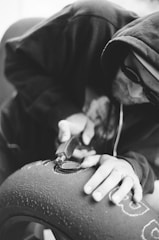 A person in a hoodie and sunglasses is engaged in an intricate work on a tire, using a tool to carve or engrave details. The focus is on the hands and the tool in action, highlighting concentration and craftsmanship.