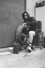 A person wearing a hoodie and sunglasses is sitting in an office chair while working on a large tire with the word 'BRUTAL' written on it. Various tools and wires are scattered around, and a battery is placed near the tire. The environment appears industrial, with a metal door and brick flooring.