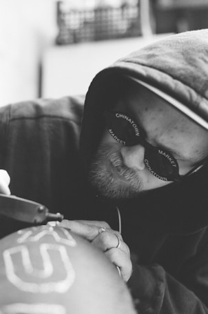 A person wearing sunglasses and a hoodie is diligently working on a craft project. The focus is on the detailed, careful application of a tool to the surface being decorated, with the word 'CHINATOWN MARKET' visible on the glasses.