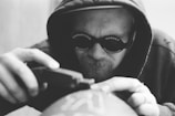 A person wearing a hood and sunglasses is intently focused on using a tool, possibly engraving or printing on a surface. The sunglasses have the words 'Chinatown Market' printed on them. The image is in black and white, emphasizing the details and textures.