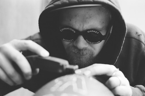 A person wearing a hood and sunglasses is intently focused on using a tool, possibly engraving or printing on a surface. The sunglasses have the words 'Chinatown Market' printed on them. The image is in black and white, emphasizing the details and textures.