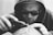A person wearing a hood and sunglasses is intently focused on using a tool, possibly engraving or printing on a surface. The sunglasses have the words 'Chinatown Market' printed on them. The image is in black and white, emphasizing the details and textures.