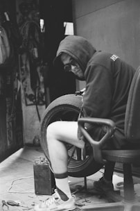 A person wearing a hoodie and sunglasses is seated on an office chair while working on a tire in a workshop. The environment is cluttered with various tools and equipment. The scene has a vintage black and white feel, emphasizing an industrial and hands-on atmosphere.