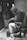 A person wearing a hoodie and sunglasses is seated on an office chair while working on a tire in a workshop. The environment is cluttered with various tools and equipment. The scene has a vintage black and white feel, emphasizing an industrial and hands-on atmosphere.