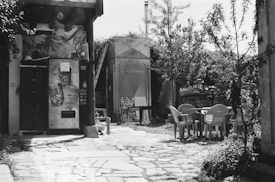 A black and white image depicts an outdoor setting with a vending machine on the left. Several plastic chairs and a table are arranged under trees to the right. The stone-paved ground and surrounding vegetation give the area a rustic and somewhat overgrown appearance. A building with wooden beams and a mural is partially visible, and a ladder is propped against a structure in the background.