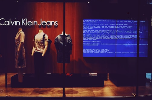A storefront display of Calvin Klein Jeans features three mannequins dressed in trendy casual wear. The background includes a large blue error screen, known as the 'blue screen of death,' commonly associated with Windows errors. The lighting is warm, highlighting the clothing.
