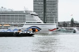 Several boats, including a large cruise ship with distinct eye graphic on its side, are in the water. A modern high-rise building looms in the background, alongside other urban structures. The scene appears to be set in a bustling harbor or port area.