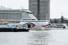 Several boats, including a large cruise ship with distinct eye graphic on its side, are in the water. A modern high-rise building looms in the background, alongside other urban structures. The scene appears to be set in a bustling harbor or port area.