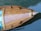 A wooden boat is seen from above, floating on calm water. The wood has a rich, polished finish with visible grain patterns. The water is a deep blue-green, with ripples reflecting light.