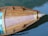 A wooden boat is seen from above, floating on calm water. The wood has a rich, polished finish with visible grain patterns. The water is a deep blue-green, with ripples reflecting light.