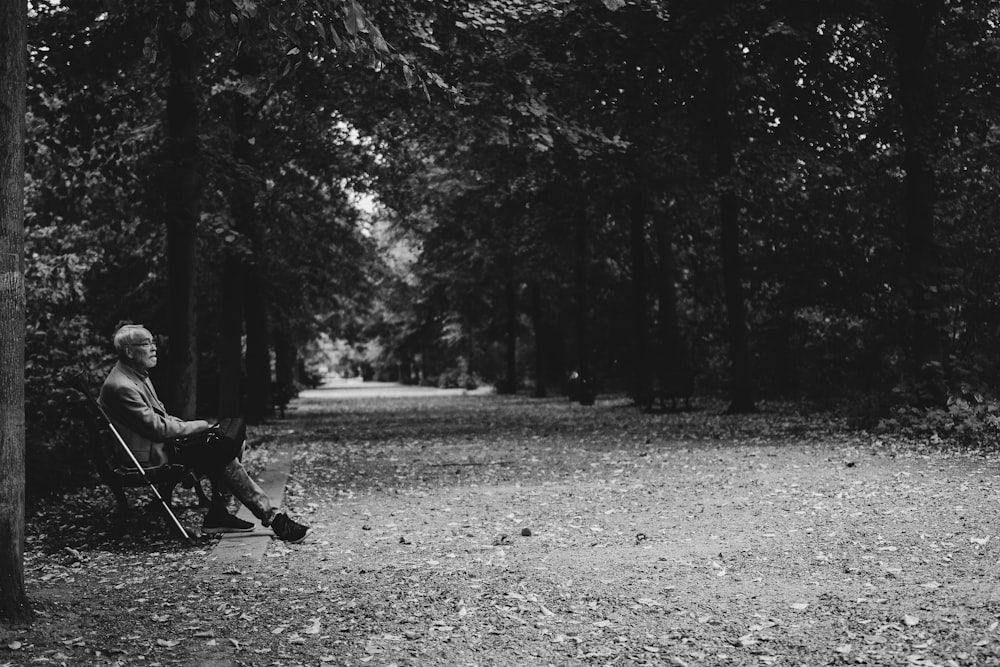 grayscale photography of old man sitting on chair near road surrounded with trees