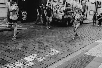 A black-and-white image of a cobblestone street in a city, featuring several people walking. One person is wearing a casual outfit with shorts, while another is in a floral dress. A parked van with advertising can be seen in the background. The street is lined with old buildings, and there are signs suggesting a European city setting.