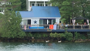 A lakeside house with a blue metal roof and a large star decoration on the wall. The deck is adorned with colorful towels and an American flag, and it features several chairs and a few people relaxing under a gazebo. Lush green trees surround the property, and two ducks swim in the tranquil water at the edge of the shore.