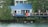 A lakeside house with a blue metal roof and a large star decoration on the wall. The deck is adorned with colorful towels and an American flag, and it features several chairs and a few people relaxing under a gazebo. Lush green trees surround the property, and two ducks swim in the tranquil water at the edge of the shore.