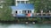 A lakeside house with a blue metal roof and a large star decoration on the wall. The deck is adorned with colorful towels and an American flag, and it features several chairs and a few people relaxing under a gazebo. Lush green trees surround the property, and two ducks swim in the tranquil water at the edge of the shore.