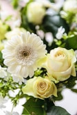 A close-up of a bouquet featuring a white gerbera daisy and pale yellow roses, surrounded by green leaves and small white flowers. The flowers are fresh and vibrant, displaying a delicate and elegant arrangement.