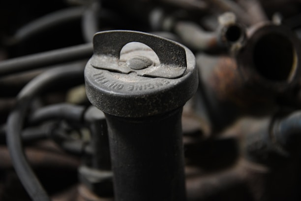 A close-up view of an old, dusty oil cap located on an engine. The cap has visible text, slightly covered with dirt and grime. Surrounding it are dark-colored hoses and metallic parts that make up the engine's components.