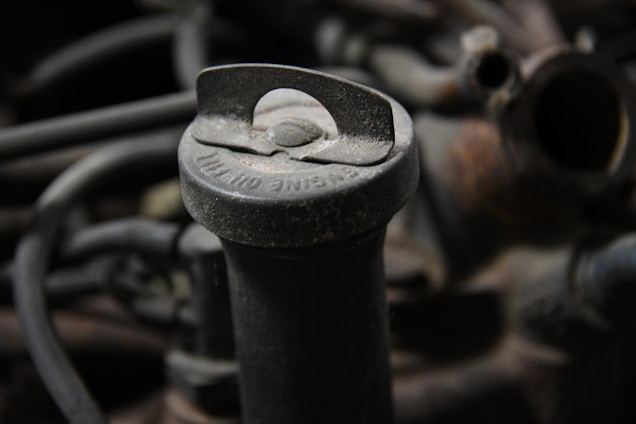 A close-up view of an old, dusty oil cap located on an engine. The cap has visible text, slightly covered with dirt and grime. Surrounding it are dark-colored hoses and metallic parts that make up the engine's components.