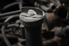 A close-up view of an old, dusty oil cap located on an engine. The cap has visible text, slightly covered with dirt and grime. Surrounding it are dark-colored hoses and metallic parts that make up the engine's components.