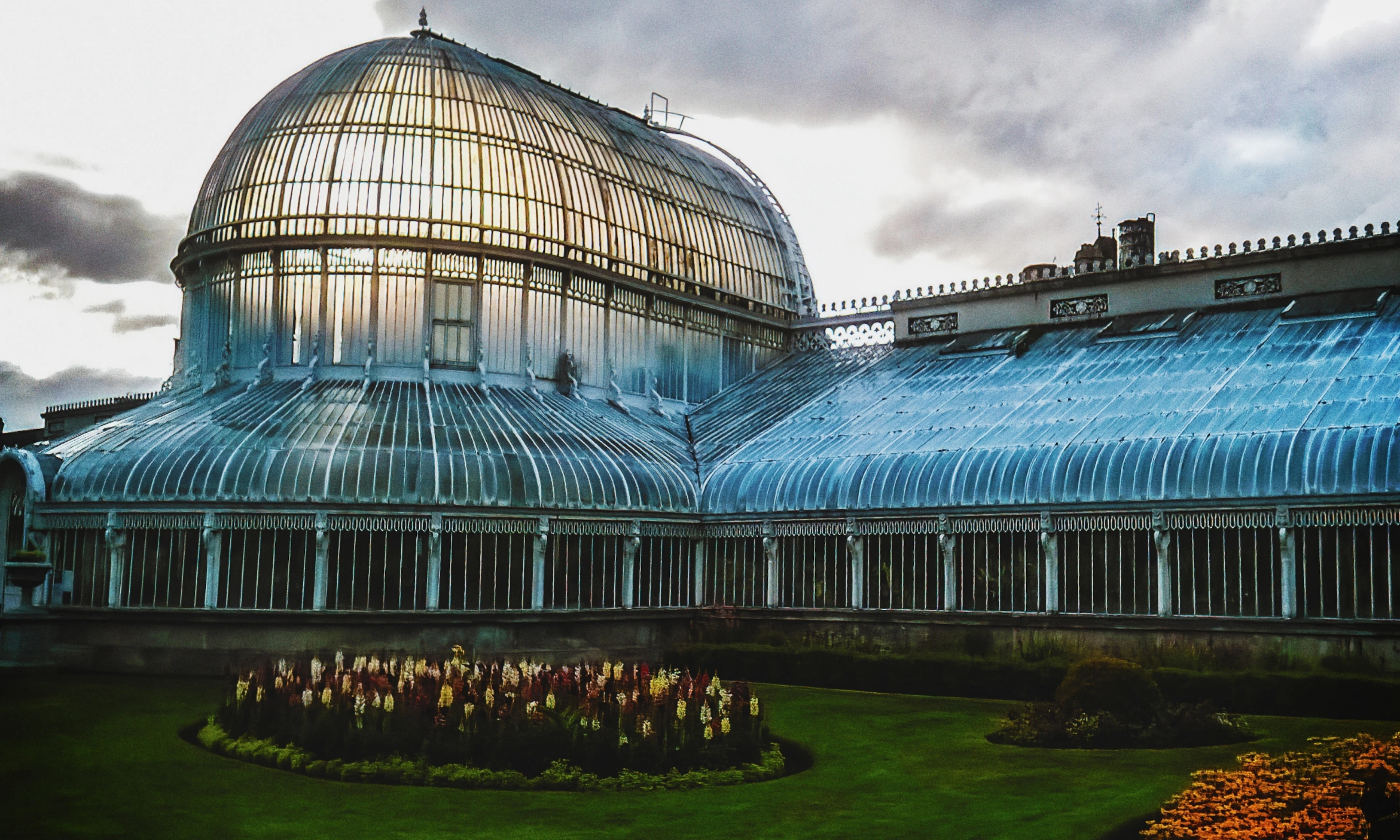 The glass-domed Palm House at Belfast's Botanic Gardens lit up at dusk with flower beds in front.