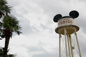 A large water tower designed with mouse ears is prominent against a cloudy sky. The tower bears the text 'Walt Disney Studios' and is situated near a palm tree, adding a touch of greenery to the scene.