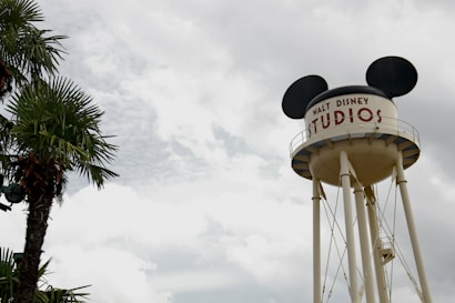 A large water tower designed with mouse ears is prominent against a cloudy sky. The tower bears the text 'Walt Disney Studios' and is situated near a palm tree, adding a touch of greenery to the scene.